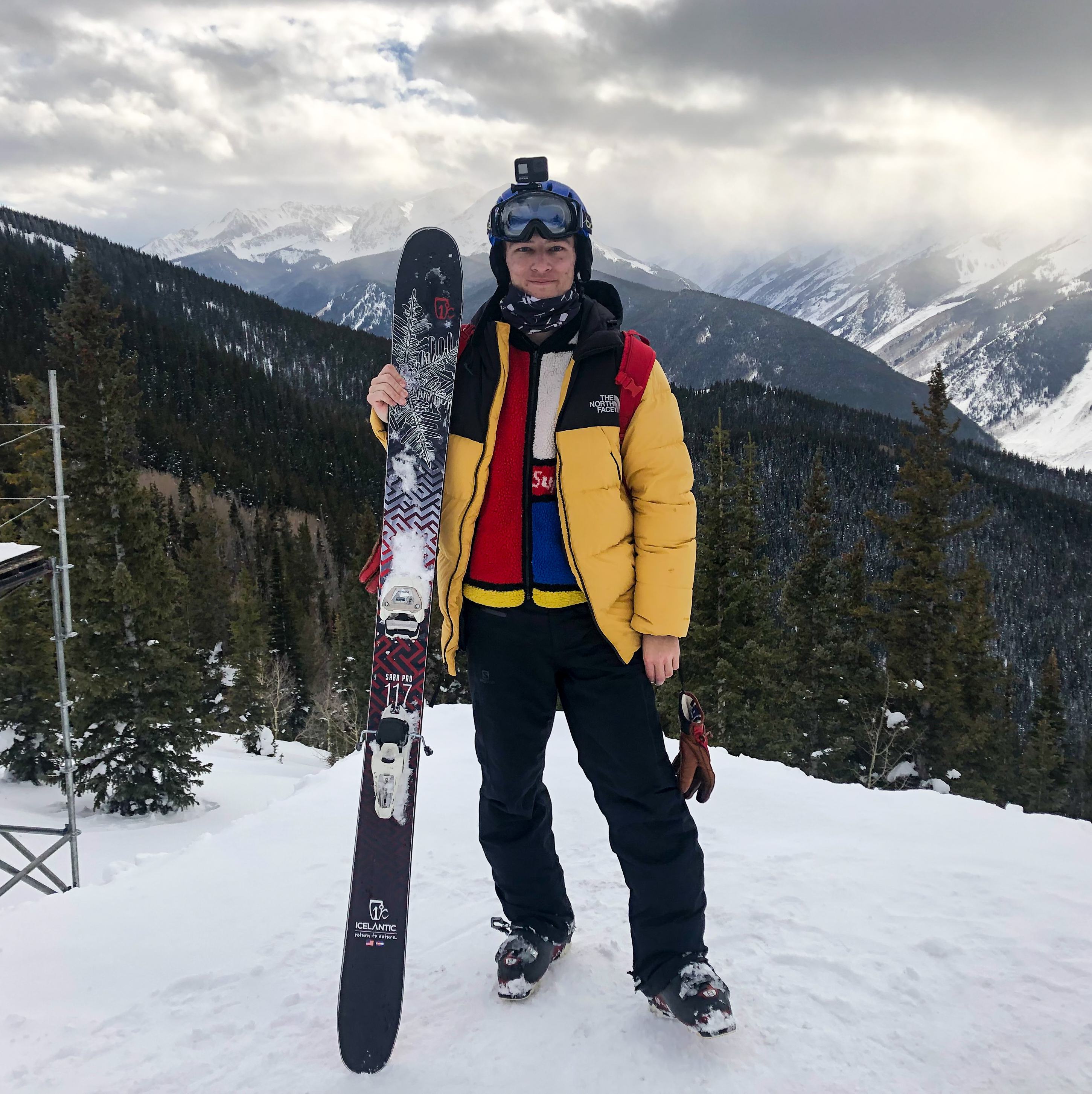 A young man standing atop a mountain with many mountains in the background. The man is wearing ski gear such as a yellow and black winter jacket, ski boots, black snow pants, red gloves, and blue helmet as well as a red backpack on his back. He is holding his large black, red, and white skis upright in his hands as he poses for the camera. His skis have a large snowflake pattern on them. 