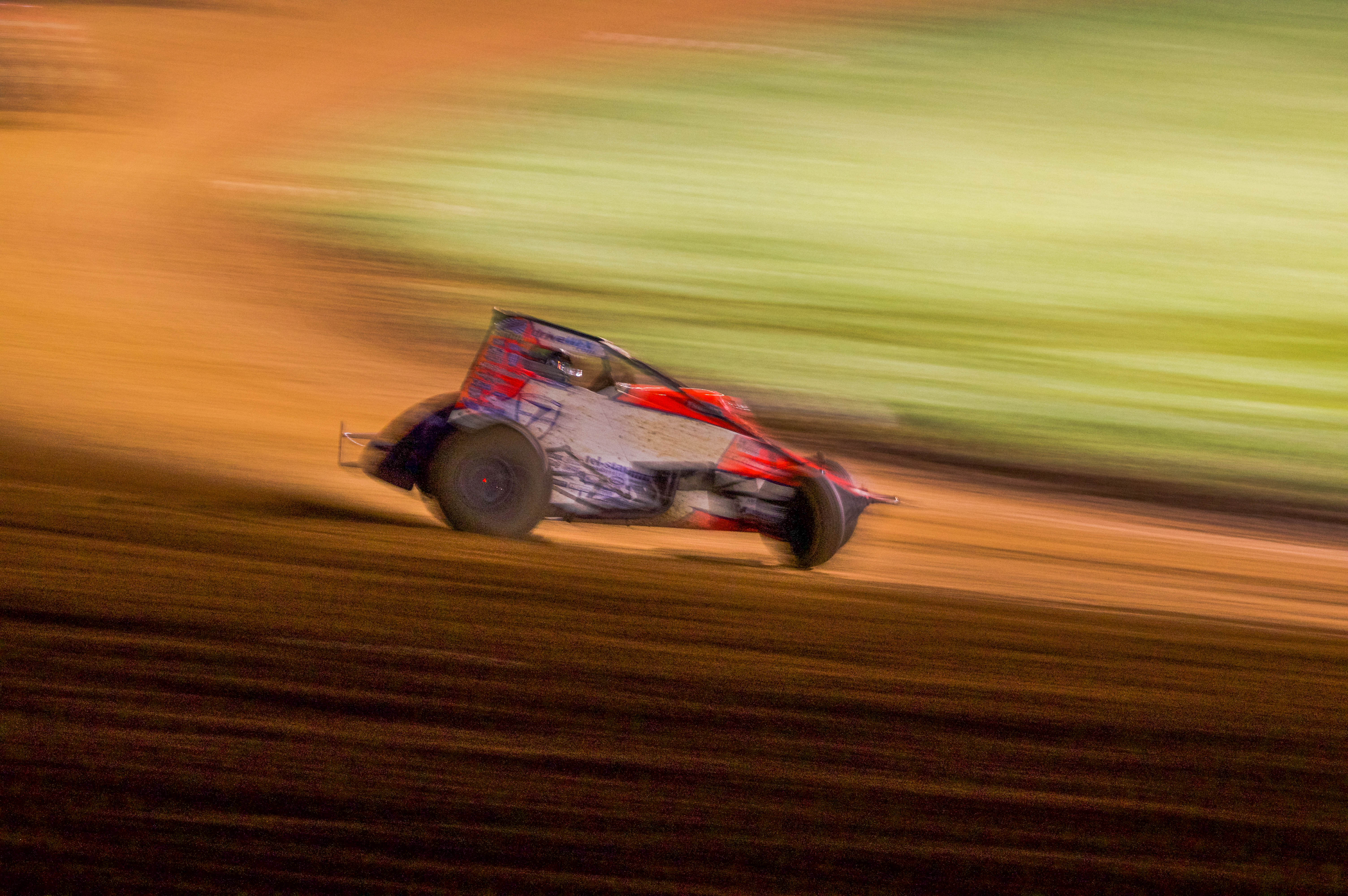 A red and blue stock car racing at high speeds at the Paragon Speedway in Bloomington, Indiana. The racecar is racing at high speeds in the night time, therefore, it is illuminated by the bright lights