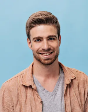 Portrait of a smiling man with light brown hair, wearing a gray t-shirt and an orange shirt, against a light blue background.