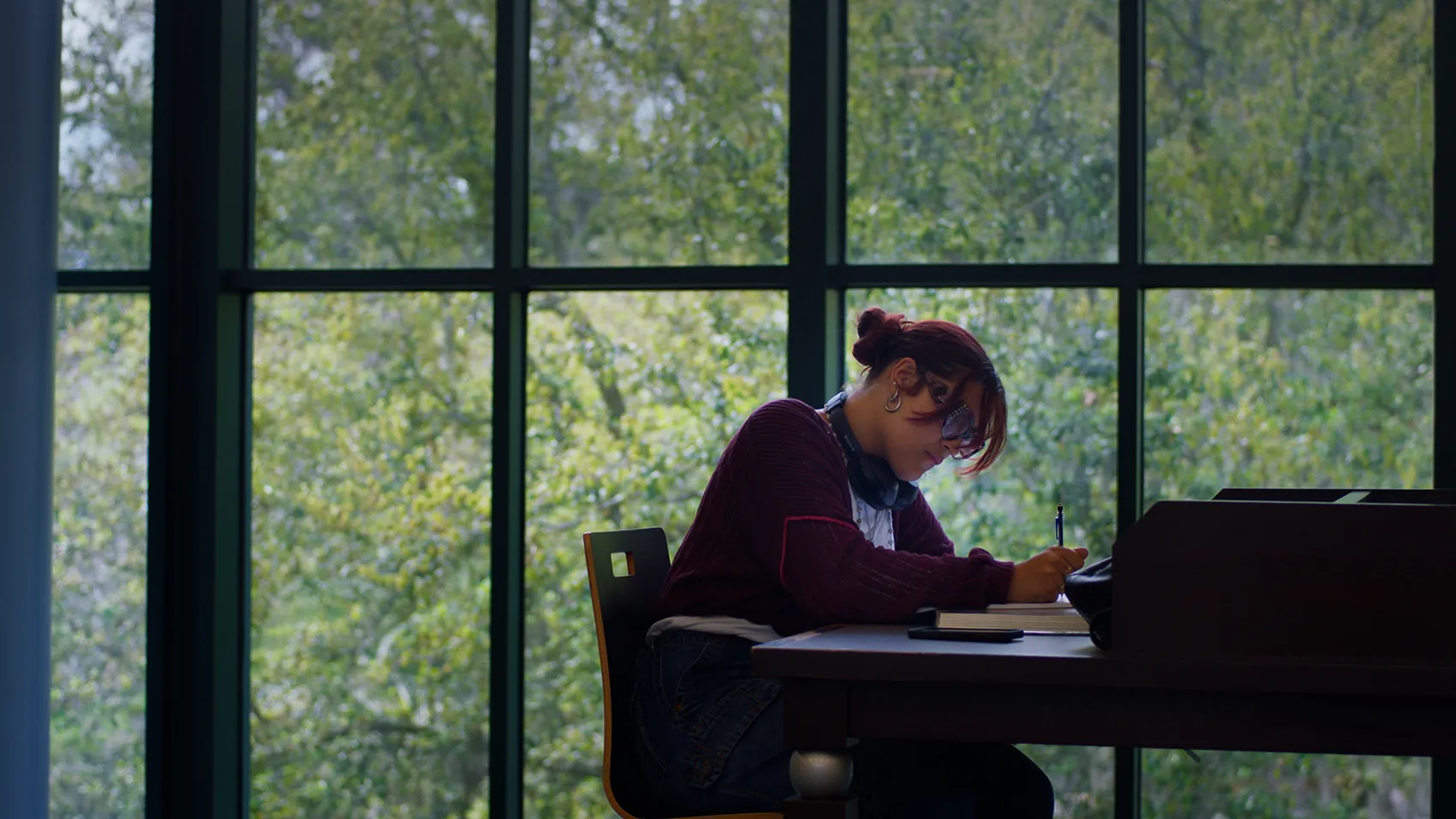 Nadya sitting at a table writing on a paper. Windows with greens or garden visible outside