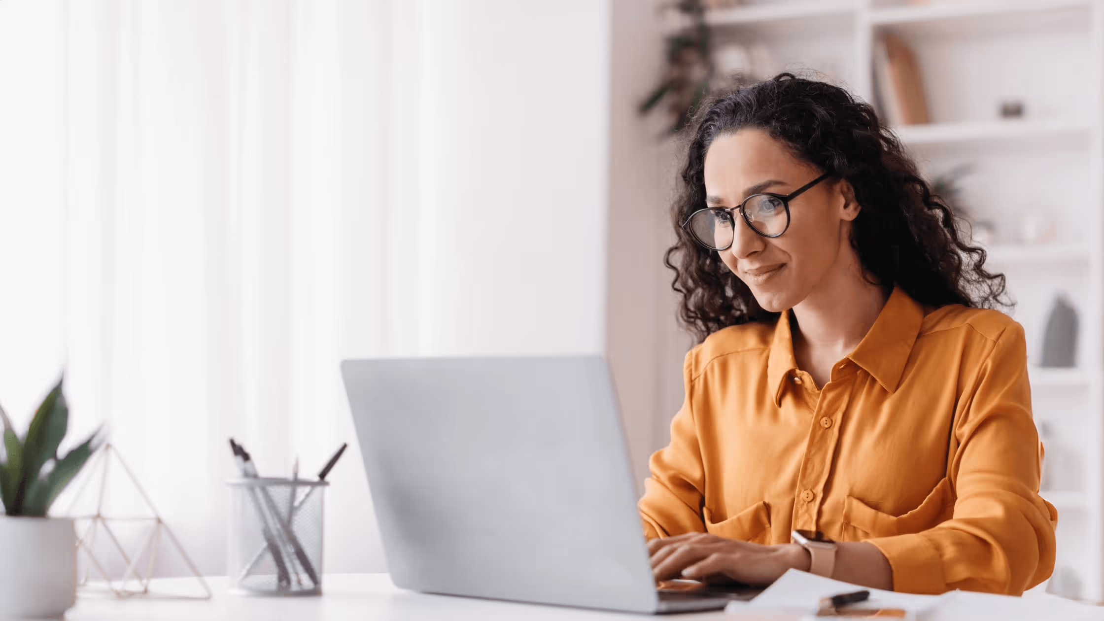 Frau mit Brille und orangefarbener Bluse arbeitet am Laptop an einem hellen Schreibtisch.