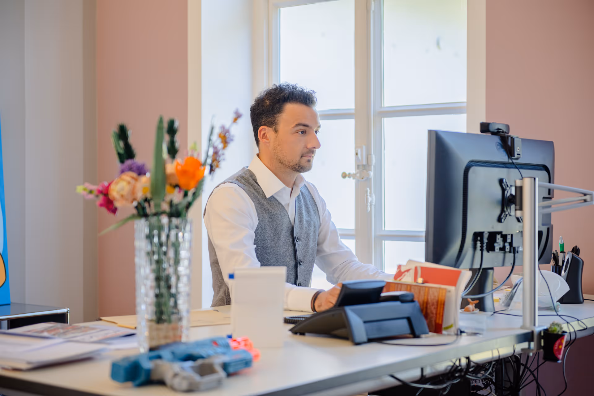 Mann in Hemd und Weste arbeitet am Computer an einem Schreibtisch mit Blumen und Büchern.