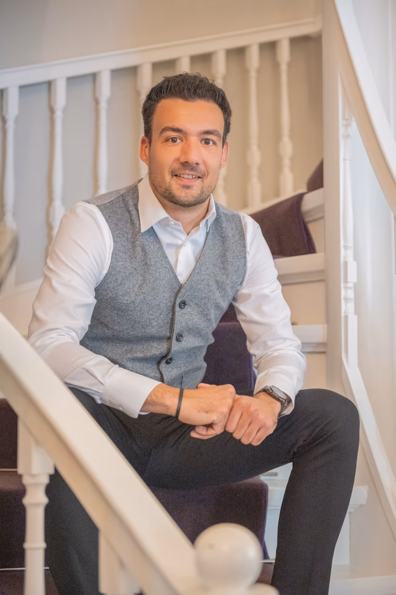 Junger Mann mit grauer Weste und weißem Hemd sitzt auf einer Treppe und lächelt in die Kamera.