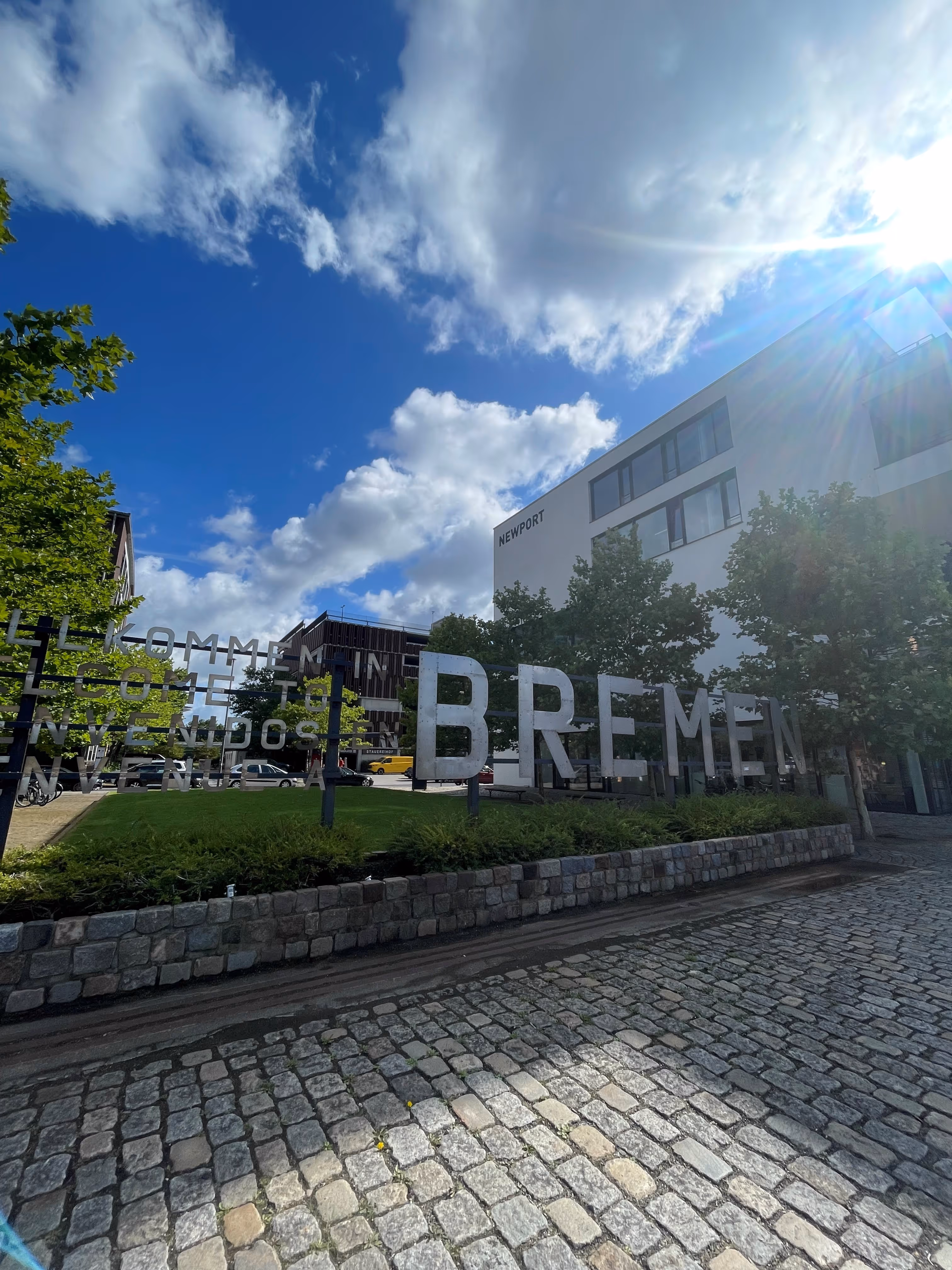 Großer Metallschriftzug 'BREMEN' vor modernem Gebäude mit blauem Himmel und Wolken.