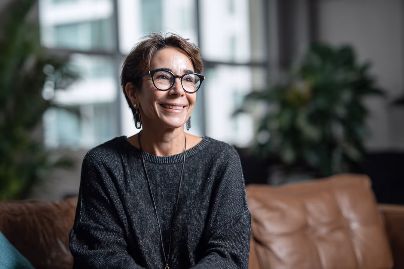 Lächelnde Frau mit Brille und dunklem Pullover sitzt auf braunem Sofa in modernem Raum.
