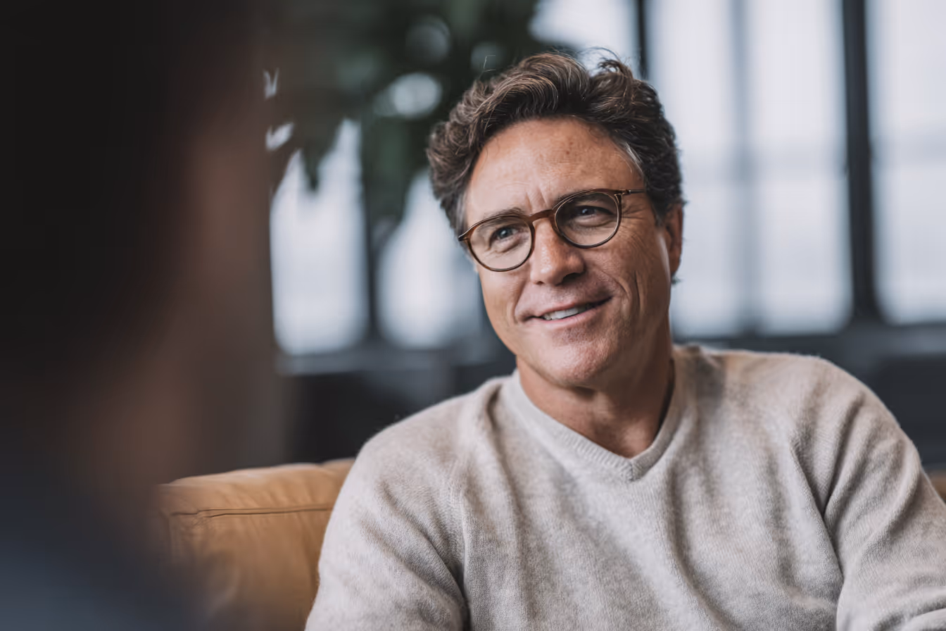 Mann mit Brille und grauem Pullover lächelt in einem modernen, hellen Innenraum.