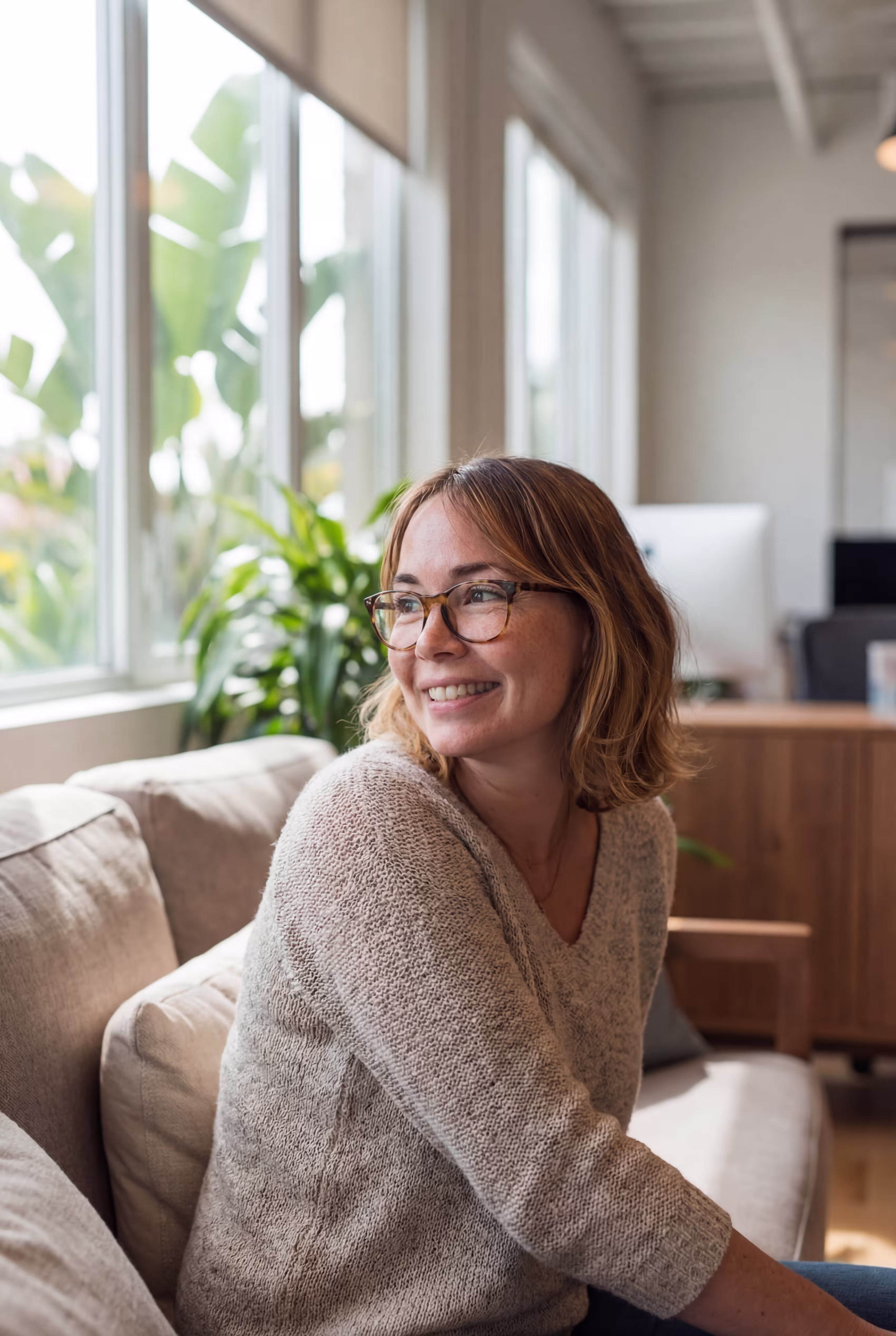 Frau mit Brille und beigem Pullover sitzt lächelnd auf einem Sofa in einem hellen Raum.