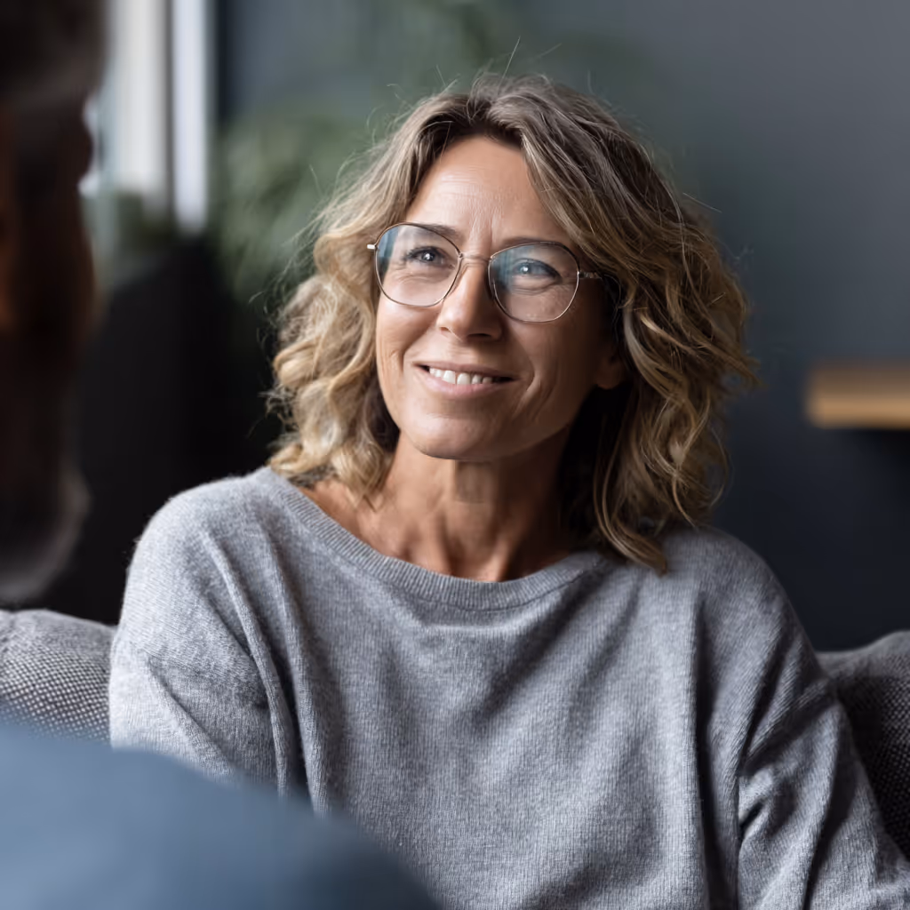 Lächelnde Frau mit lockigem Haar und Brille trägt grauen Pullover und sieht jemanden an.