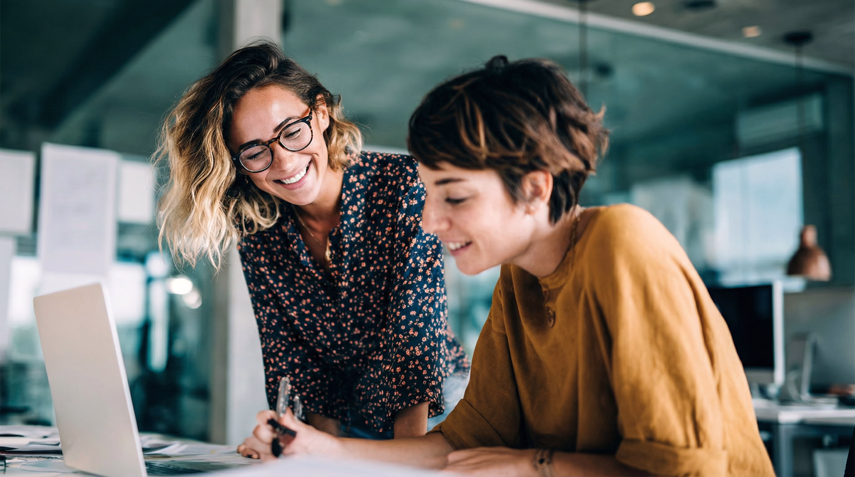 Zwei Frauen arbeiten in einem modernen Büro zusammen und lächeln über Laptop und Papier.