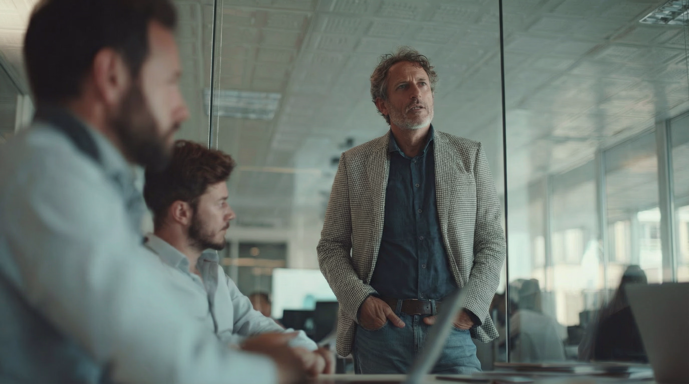 Mann in Jacke spricht in einem modernen Büro mit zwei Männern am Tisch.