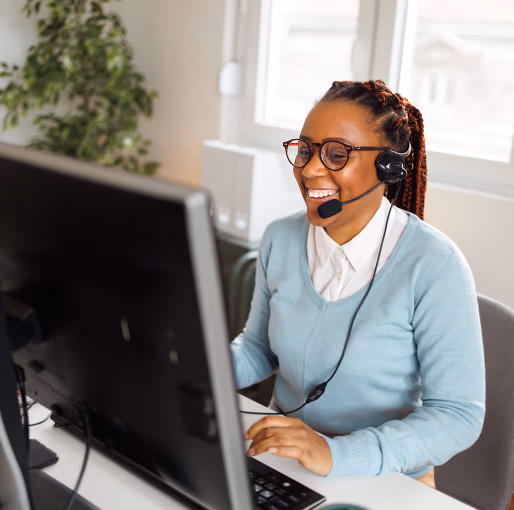 Smiling woman with glasses and headset working at a computer in a bright office, representing payers and health plans.