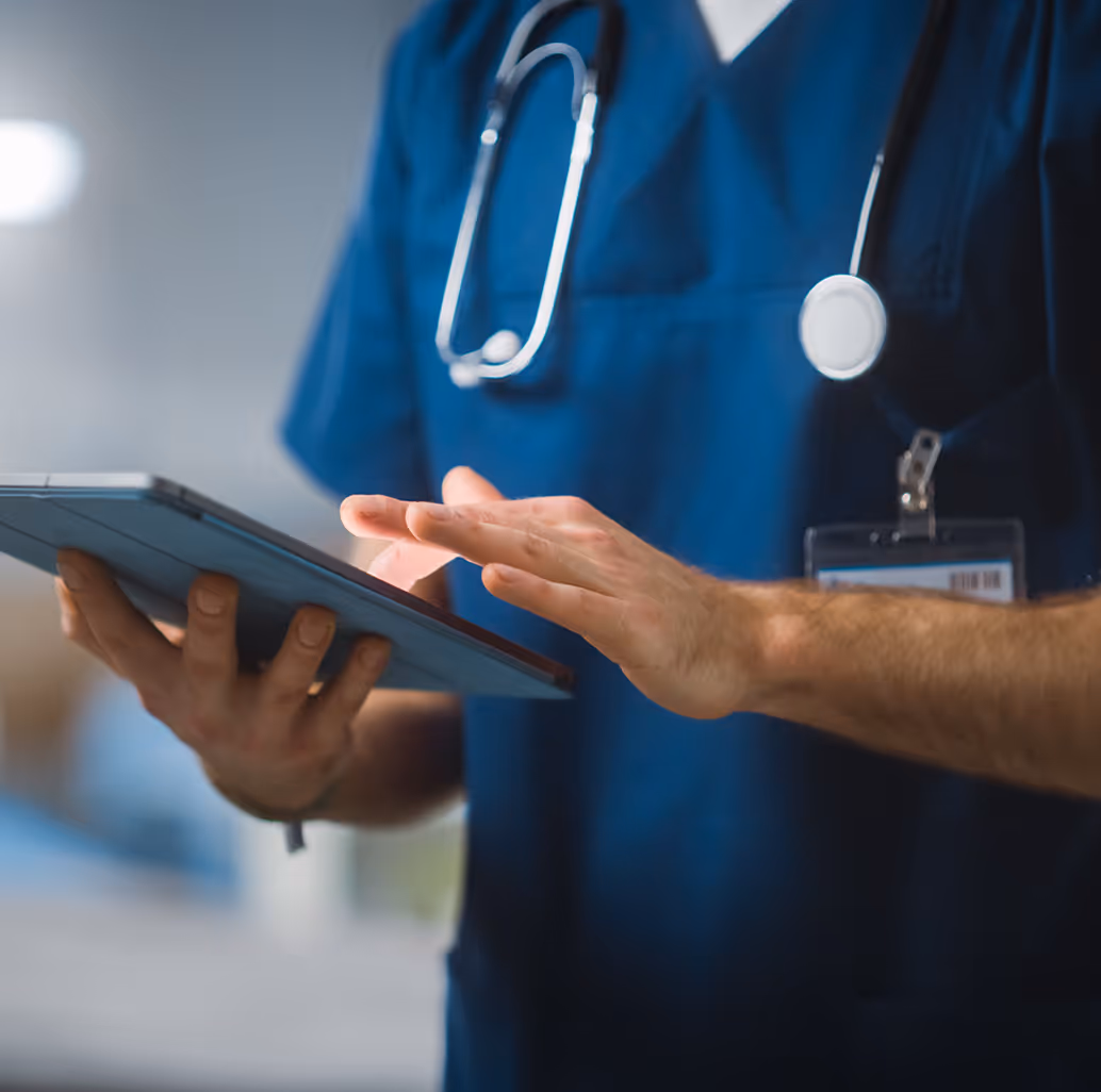 Healthcare professional in blue scrubs using a digital tablet, with a stethoscope around their neck.
