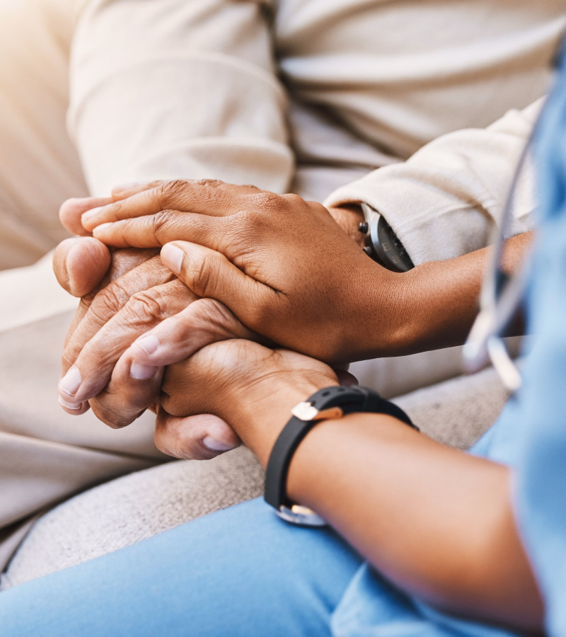 A home health nurse in blue scrubs interacts warmly with an elderly patient seated on a couch in a cozy home setting, conveying compassion and personalized care.
