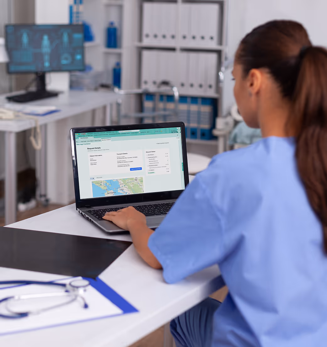 Healthcare worker in blue scrubs using a laptop showing patient transport details and status in a medical office.
