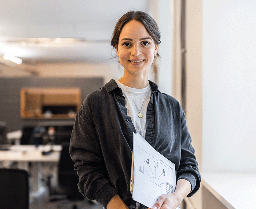 Junge Frau im Büro hält Unterlagen in der Hand und lächelt in die Kamera – symbolisiert Informationssuche zu duales Studium Kosten.