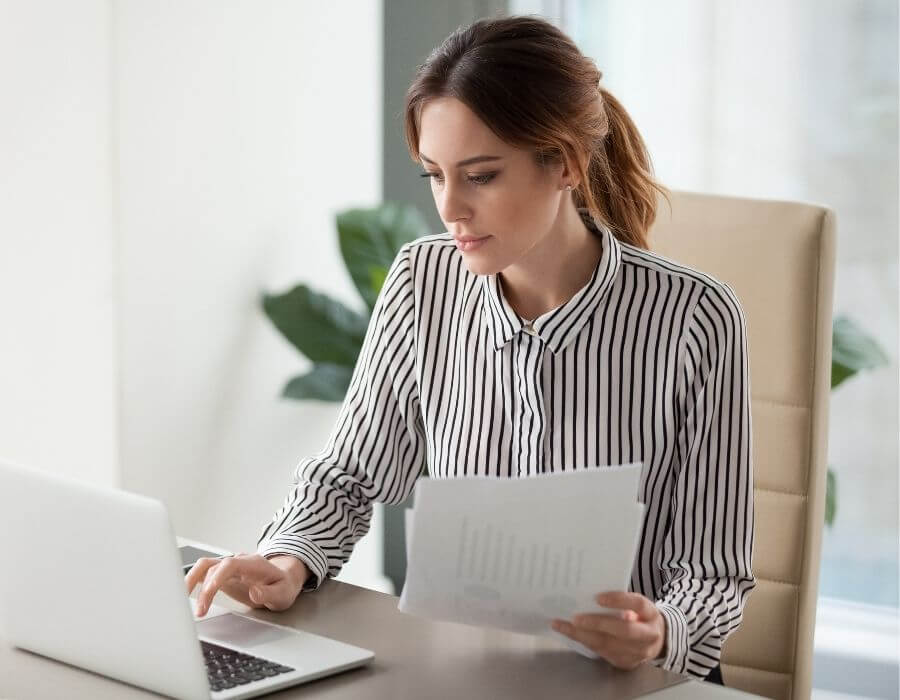 Junge Frau arbeitet konzentriert am Laptop und überprüft Unterlagen – symbolisiert die Erstellung eines Anschreibens fürs duale Studium.