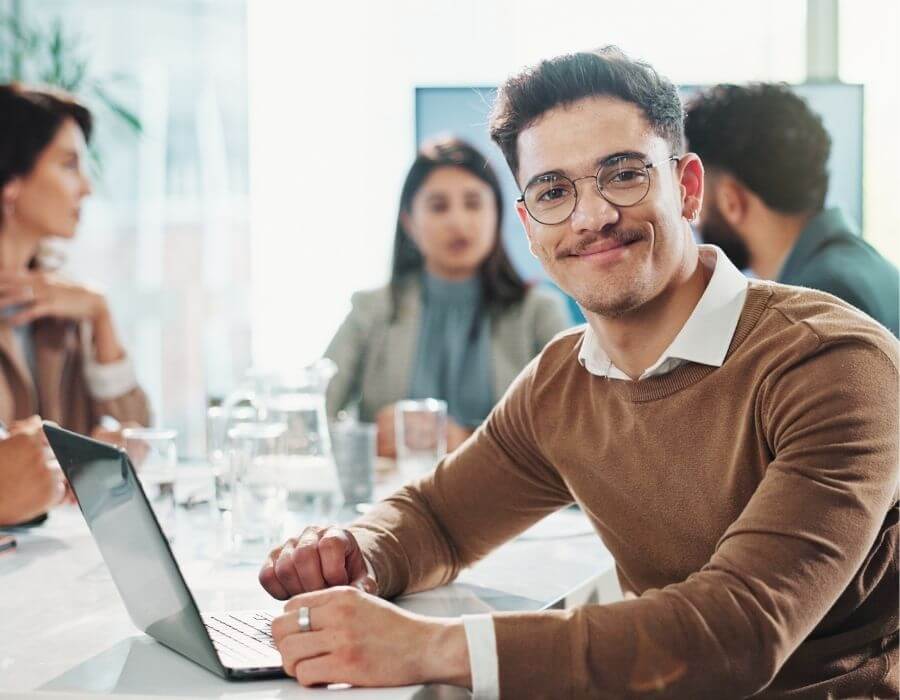 Junger Mann im Meeting mit Laptop, lächelt in die Kamera – typische Szene aus dem Alltag im dualen Studium. Vorteile eines dualen Studiums: Praxisnähe und direkte Anwendung des Gelernten im Unternehmen.