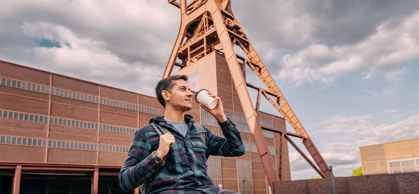 Student mit Kaffeebecher vor der Zeche Zollverein in Essen – Symbol für NRW und perfekter Ausgangspunkt für das duale Studium Soziale Arbeit in Nordrhein-Westfalen an der CBS.