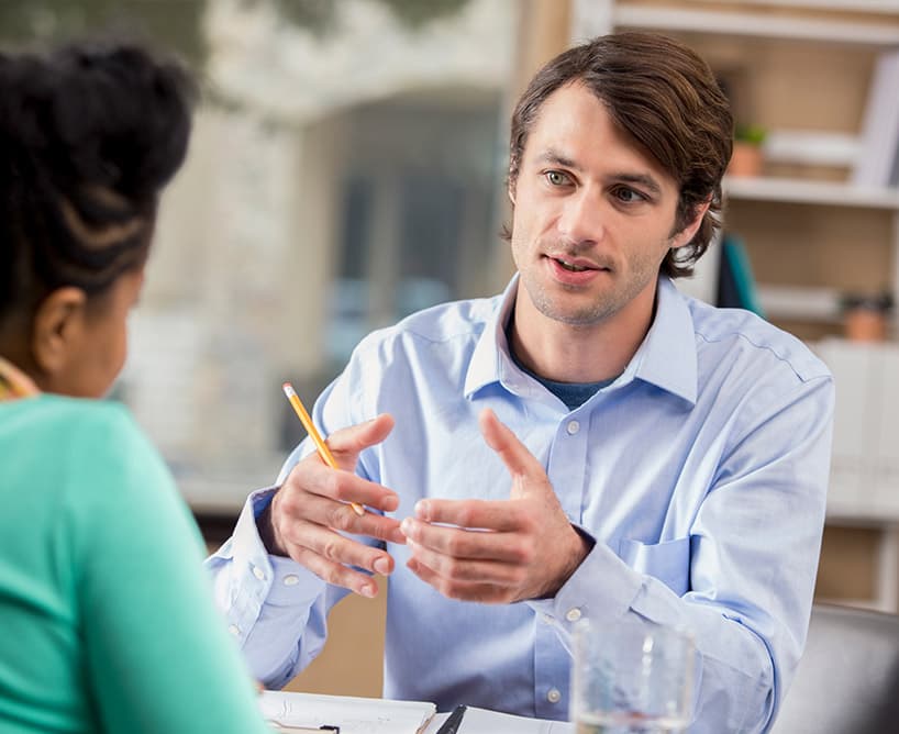 Mann im hellblauen Hemd erklärt einer Frau etwas während eines Gesprächs in einem Büro – symbolisiert Informationssuche zu duales Studium Kosten.
