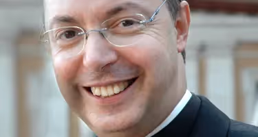 Smiling person associated with Festival di Spoleto, wearing glasses and clerical attire