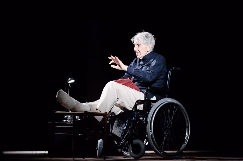 Elder performer on stage at Festival di Spoleto, seated in wheelchair, gesturing dramatically