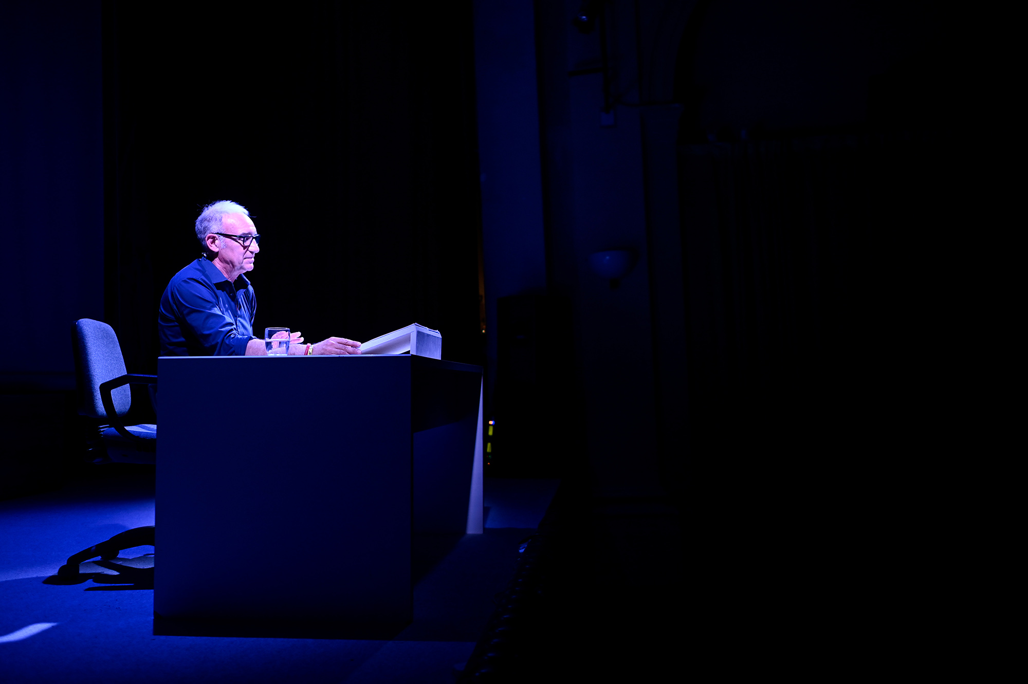 Speaker at Festival di Spoleto, seated alone on dimly lit blue stage