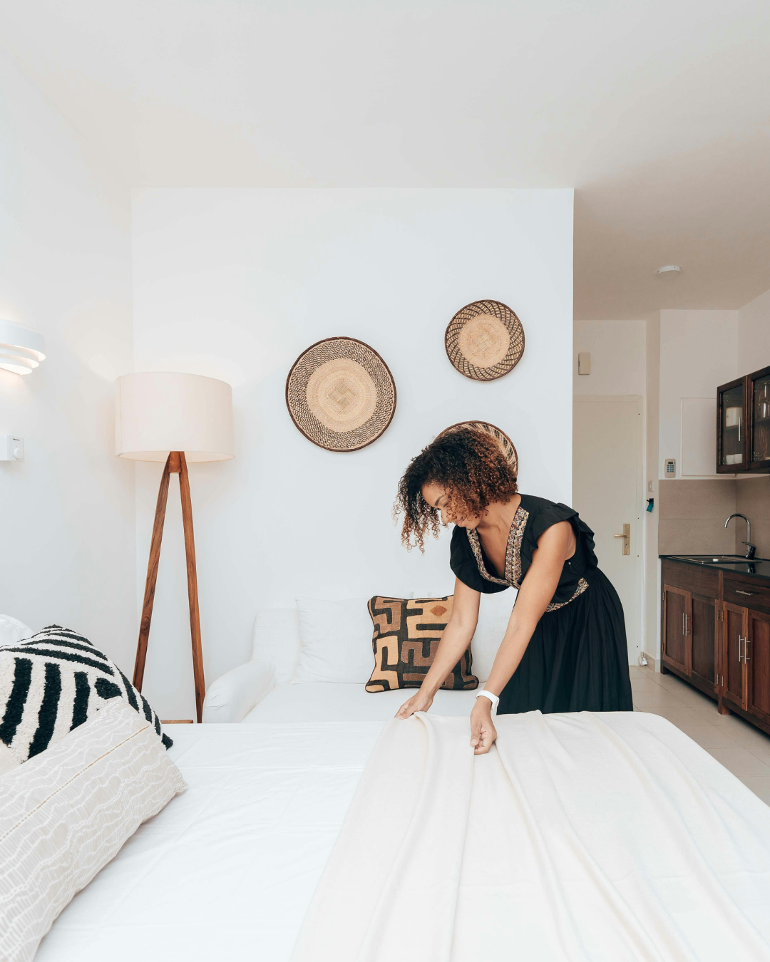Woman in black dress making a bed in a modern room with wooden lamp and woven wall decor.