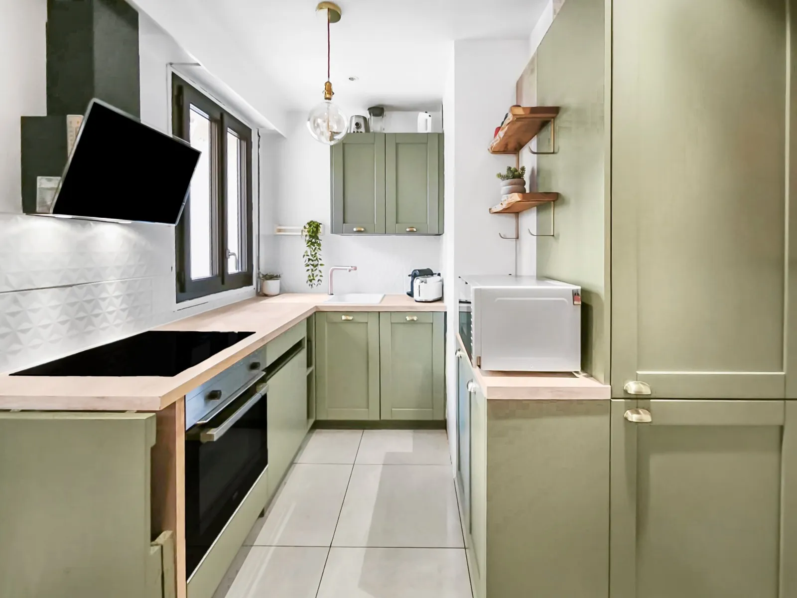 Cuisine moderne en U avec des armoires vertes, un plan de travail en bois clair, un four encastré, une hotte inclinée, un micro-ondes et une fenêtre noire.