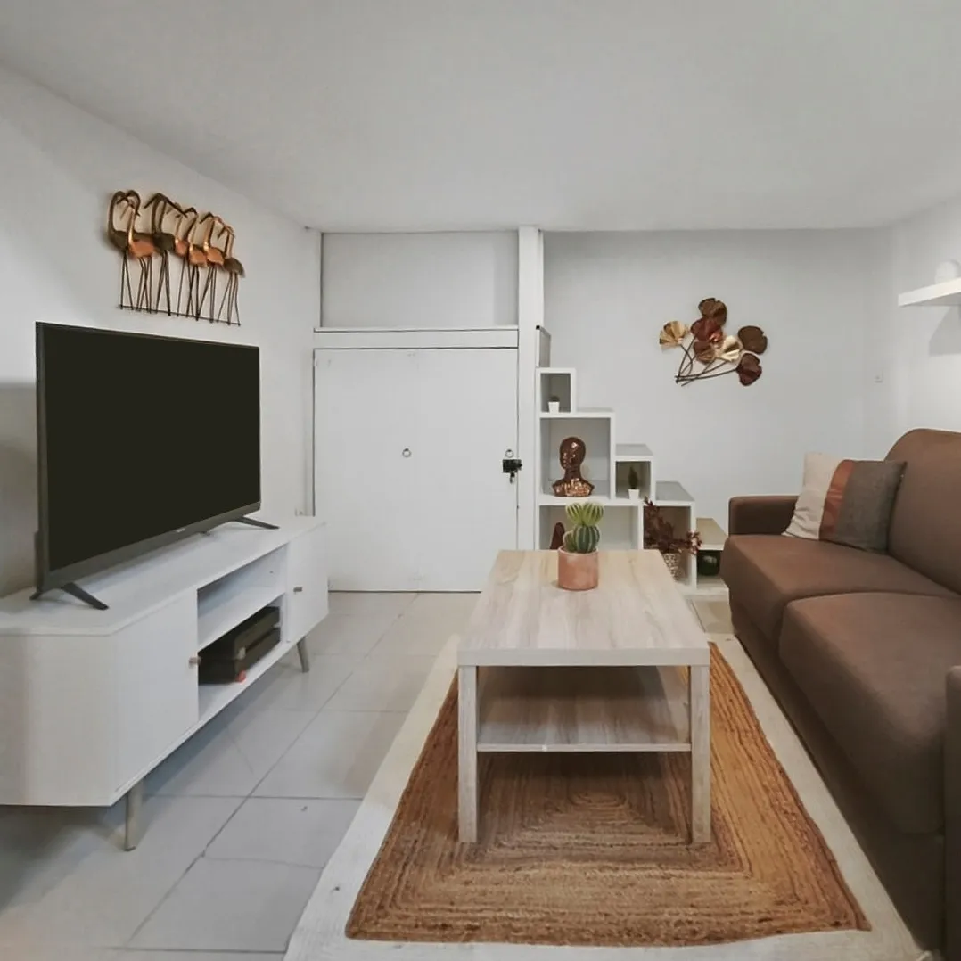 Salon moderne avec canapé marron, table basse en bois clair, tapis beige et meuble TV blanc avec télévision.
