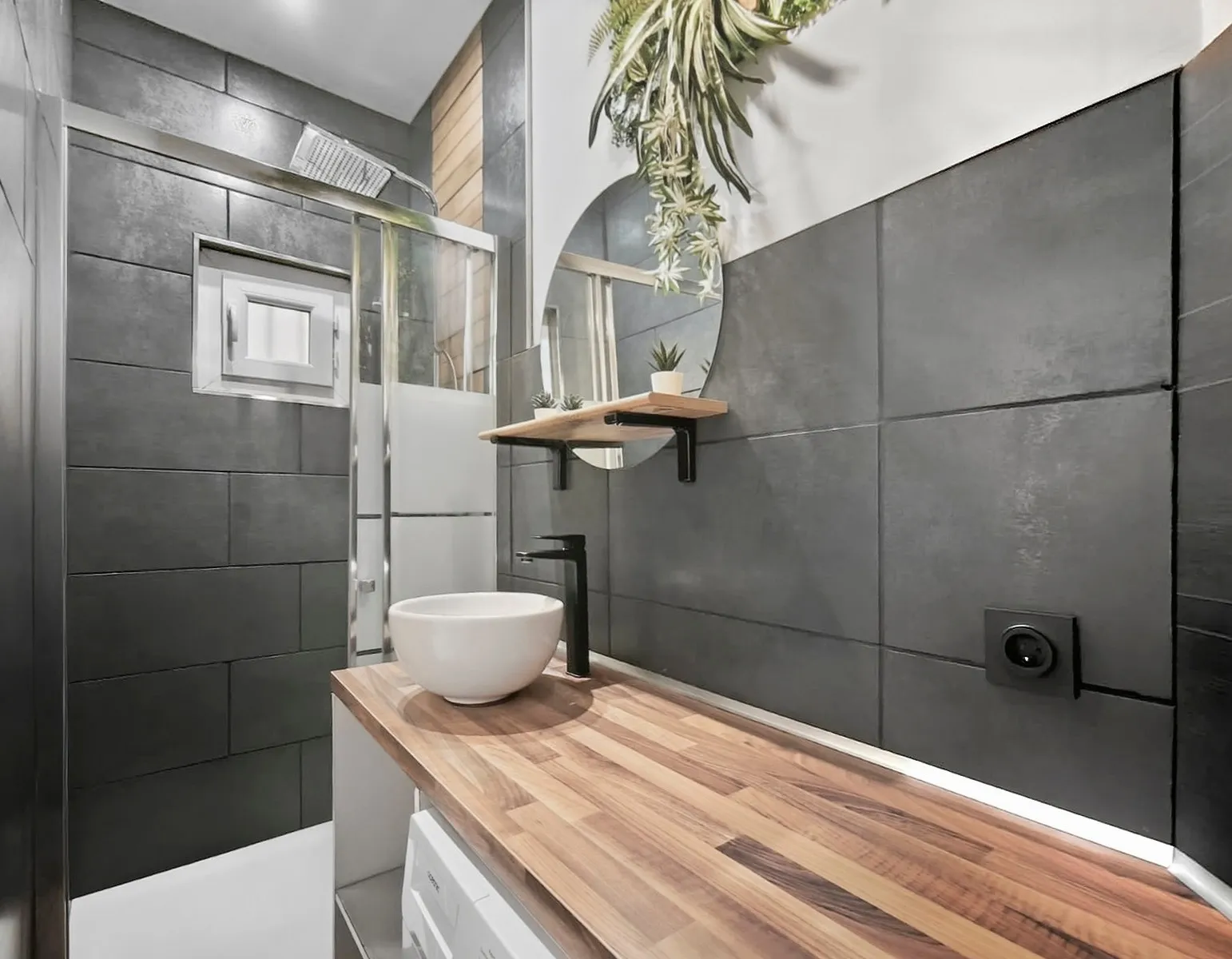 Salle de bain moderne avec carrelage gris foncé, lavabo blanc rond sur un plan de travail en bois et une douche avec paroi en verre.