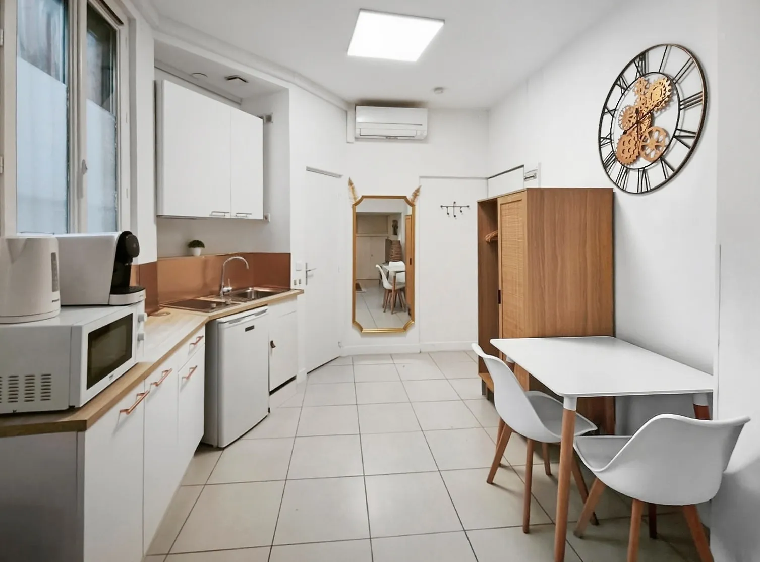 Cuisine moderne avec carrelage clair, comptoir en bois, micro-ondes, petite table blanche avec deux chaises, armoire en bois et une horloge murale décorative.