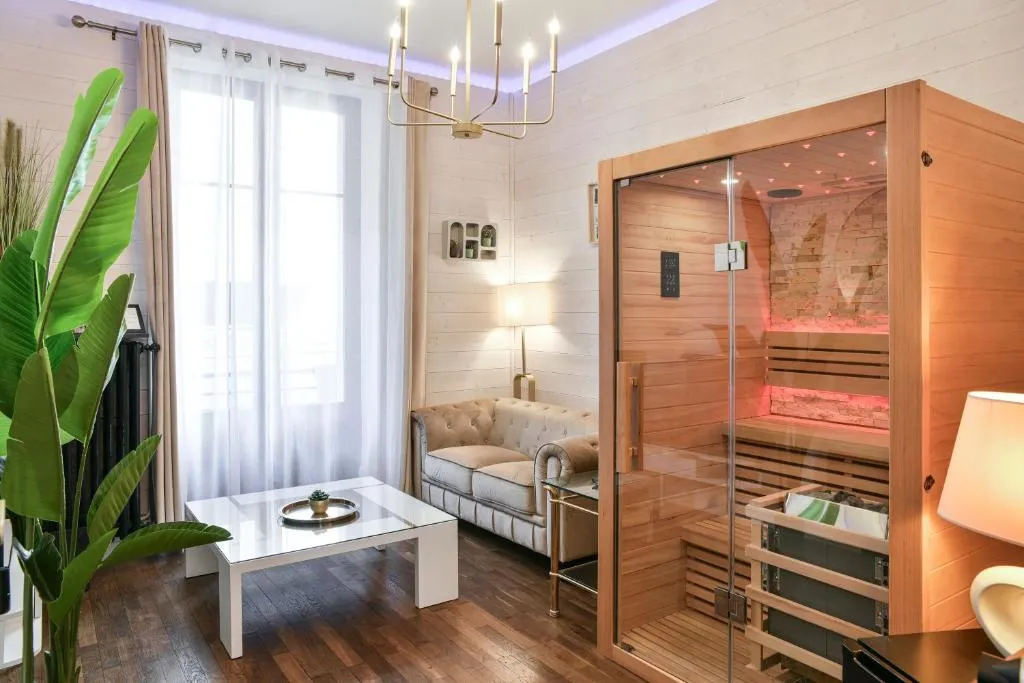 Salon moderne avec canapé beige, table basse blanche, grande plante verte et sauna en bois avec porte en verre.