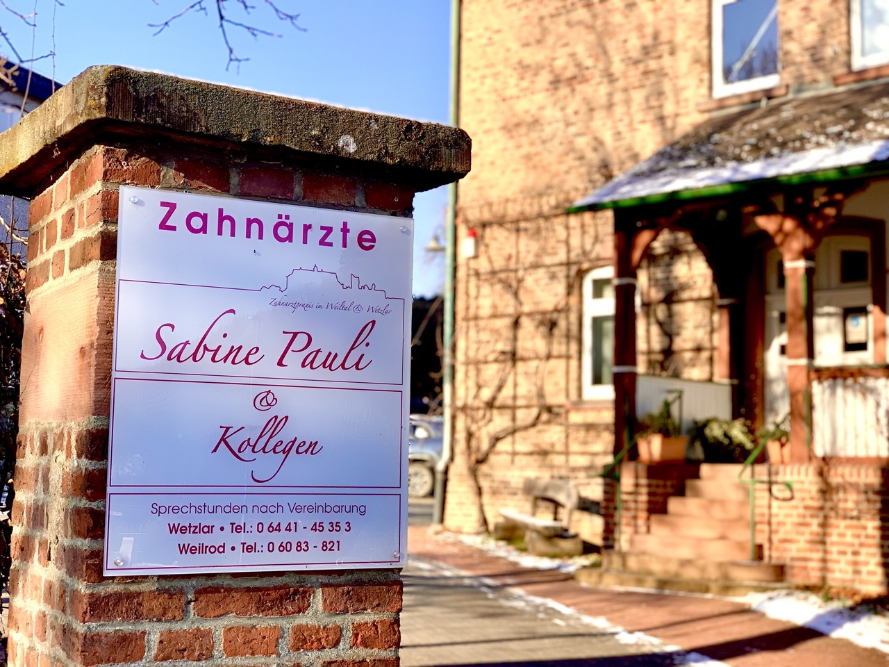 Eingang der Zahnarztpraxis mit Praxisschild „Zahnärzte Sabine Pauli & Kollegen“ an einer Backsteinmauer.