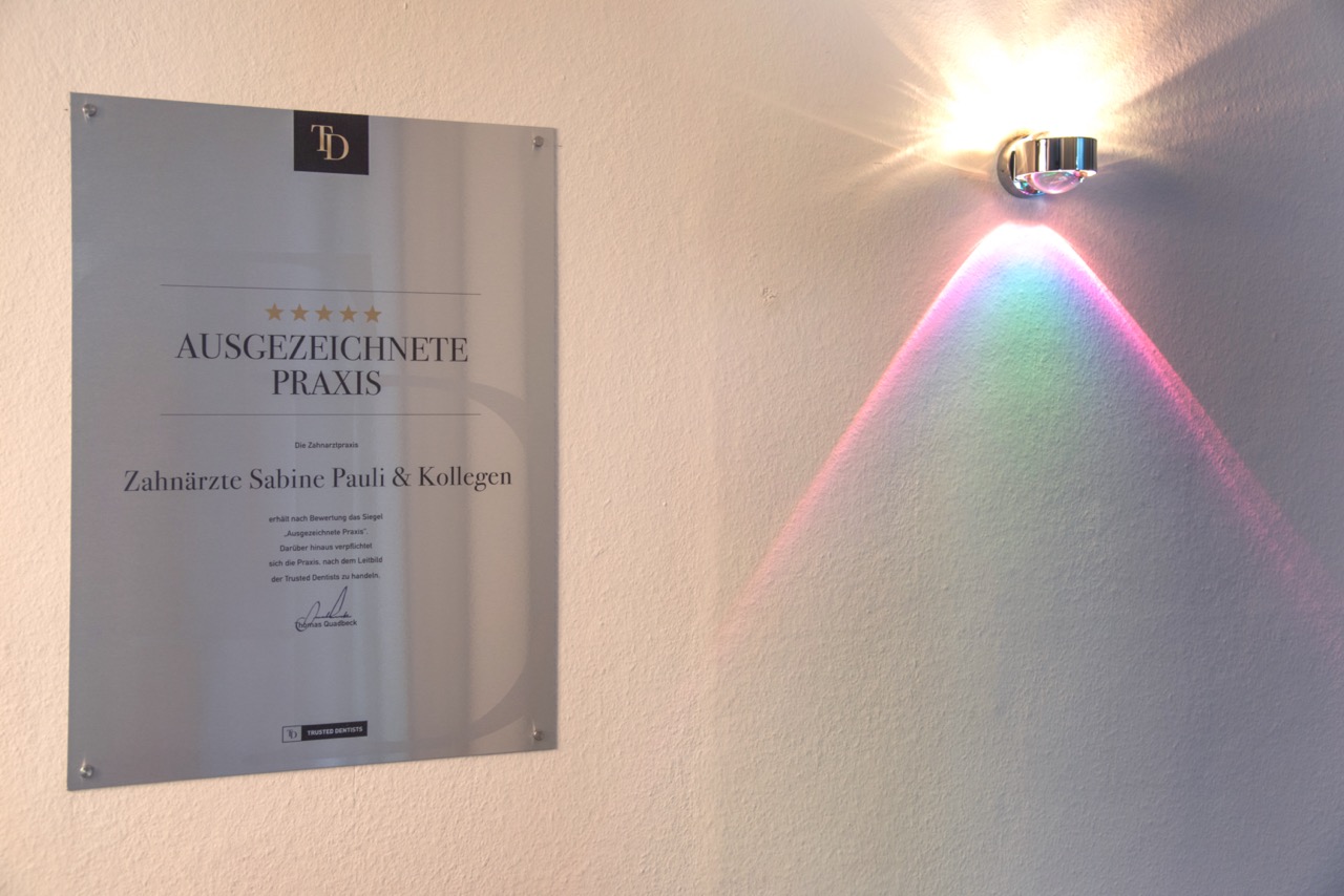 Auszeichnung an der Wand mit der Aufschrift „Ausgezeichnete Praxis“ neben einer Wandlampe mit Lichtreflexion.
