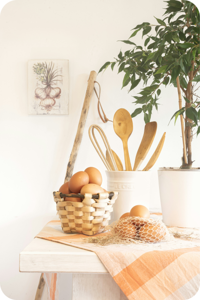 Rustic kitchen scene with eggs in a woven basket, wooden cooking utensils in a white container, a plant in a white pot, and a kitchen towel on a light wooden table.