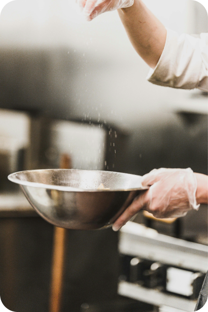 A man standing in a kitchen dropping spice into a stainless steel bowl.