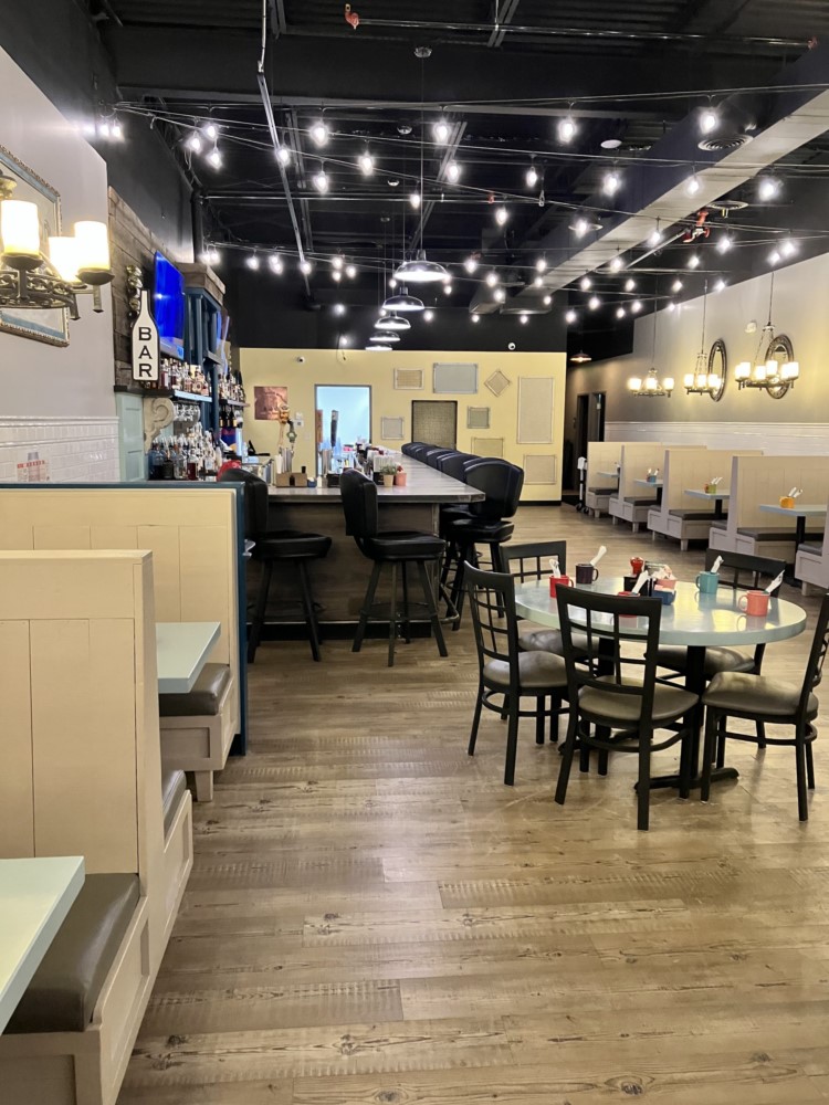 The interior of Samantha's Grill at Oakwood Square in Plain Township, OH. Displays wooden floors, beige booths along the walls, a central bar with black bar stools, round tables with chairs, and string lights hanging from the ceiling.