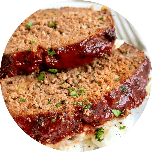 Close-up of two slices of meatloaf with a glazed sauce and sprinkled herbs on top, served on a white plate.