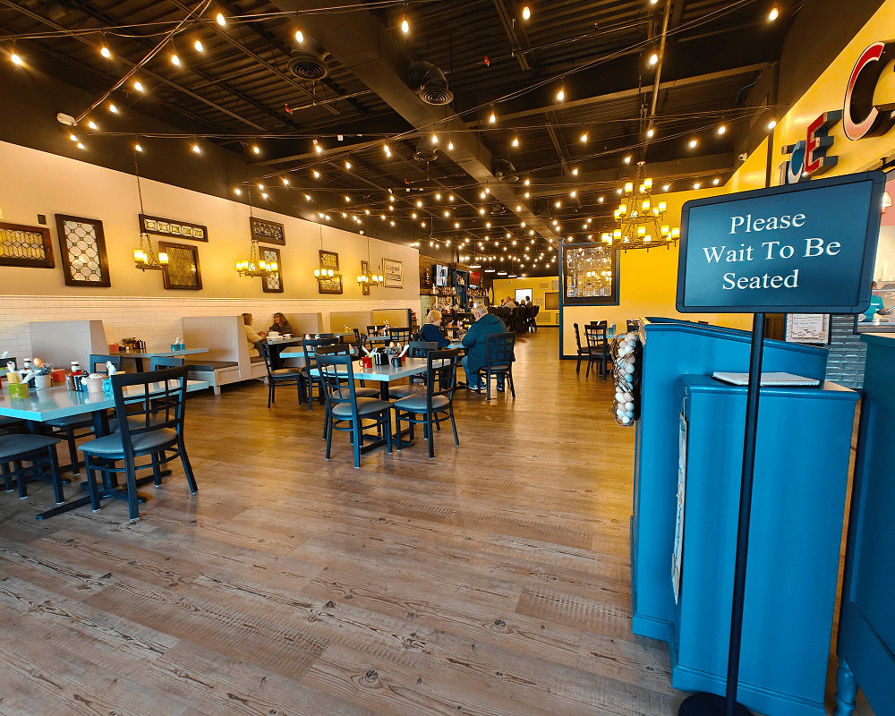 The interior of Samantha's Grill at Oakwood with wooden floor, blue booths, tables with black chairs, string lights, and a sign reading 'Please Wait To Be Seated.'
