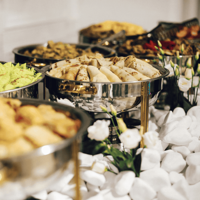 Buffet table with stainless steel chafing dishes filled with assorted folded crepes and other dishes, decorated with white flowers and stones.