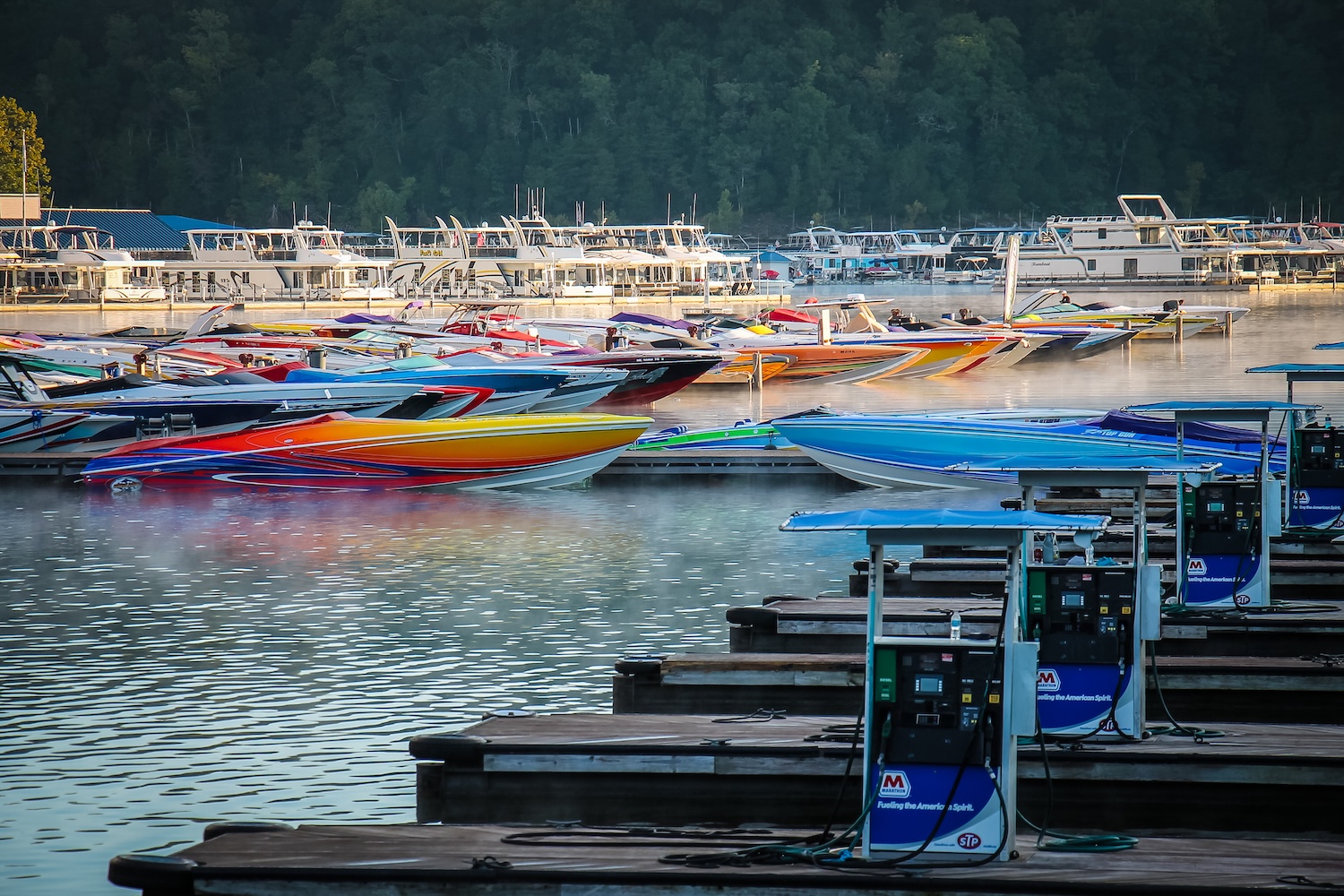 Colorful speedboats docked at a Lake Cumberland marina with calm water, fuel pumps, and forested hills in the background.