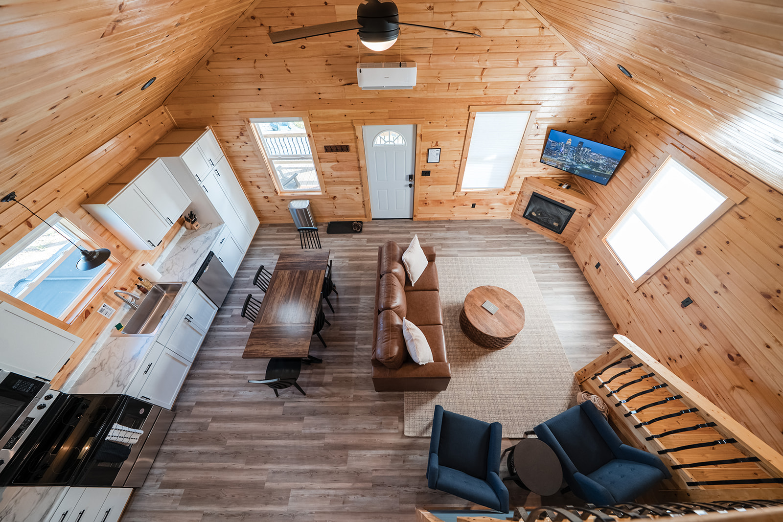 Bright modern cabin interior with wood-paneled walls, open kitchen, dining table, cozy living area, and lofted ceiling viewed from above.