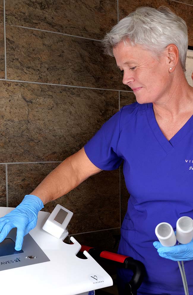 Kvinne med kort grått hår i blått medisinsk arbeidstøy og hansker bruker en medisinsk enhet i klinikk.