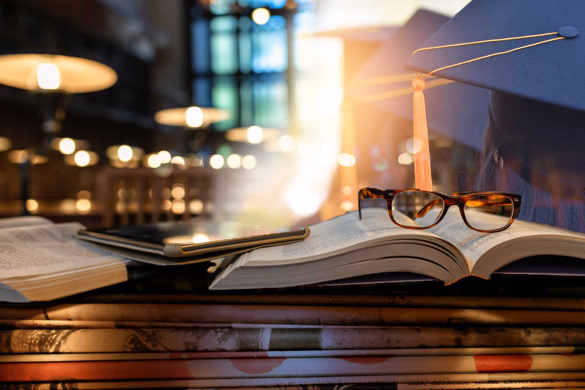 Graduation cap and glasses resting on open books with warm bokeh lighting