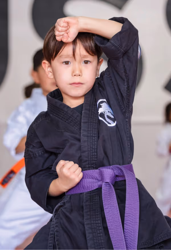A young karate student wearing a purple belt practices a block position in karate class.