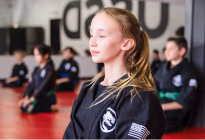 A teenage girl is meditating in a seated position in a dojo along with other participants of her teens class