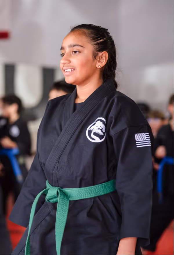 A teenager in a black karate gi and green belt is smiling while she waits during karate class.