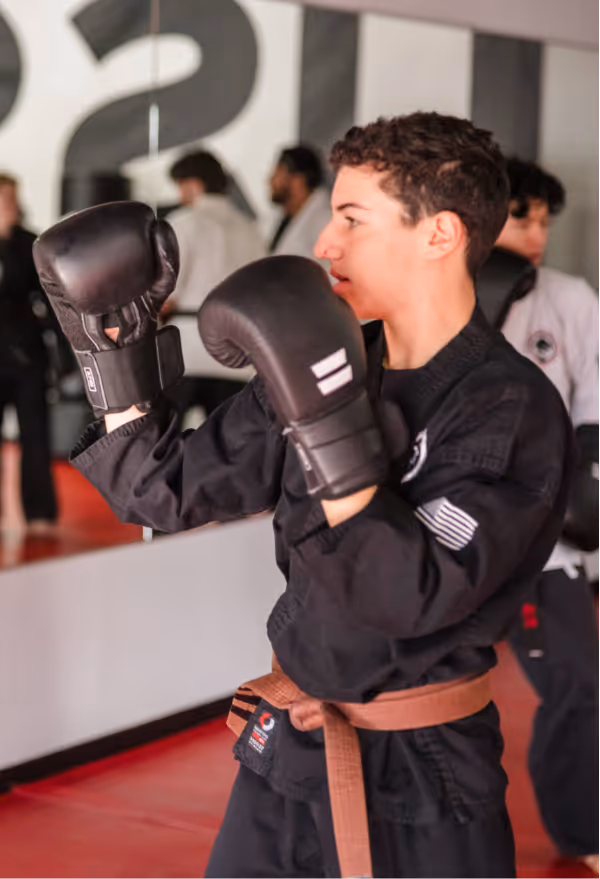 A teenager in a karate gi with a brown belt is wearing boxing gloves as he spars during karate class.