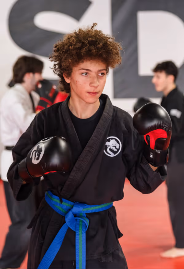 A teenager with fluffy curly hair is on guard, wearing boxing gloves and a karate gi with blue belt as they spar during karate class.