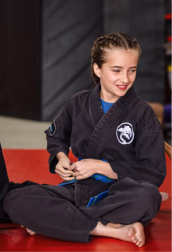 A smiling teenage girl wearing a black karate gi with a blue belt sits cross-legged on the floor with her friends to chat before karate class begins.